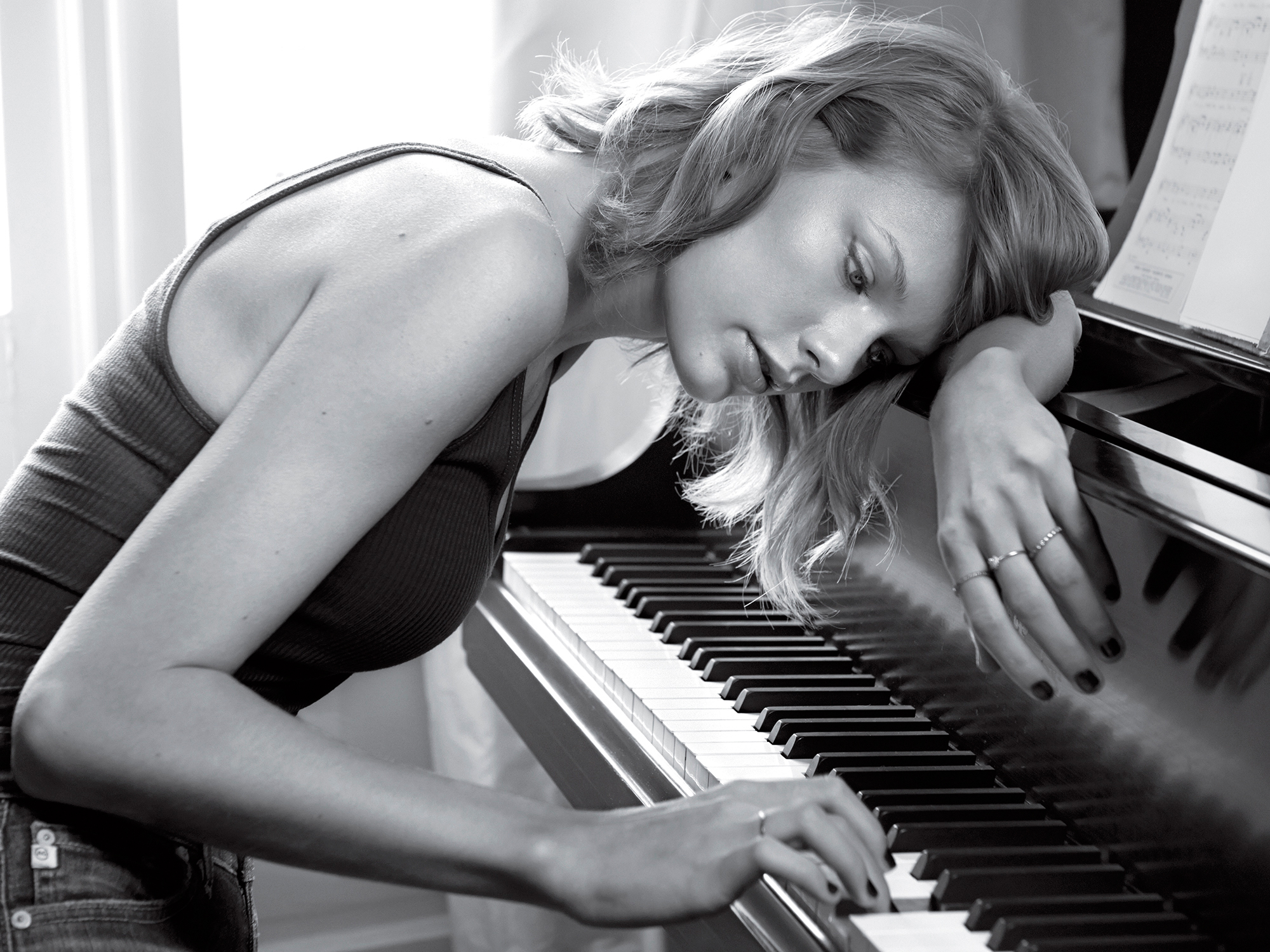 A black and white image of Taylor Swift leaning against a piano, one hand on the keys.